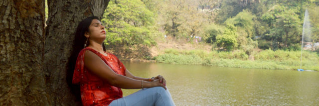 Indian woman sitting against a tree in a red shirt and blue jeans looking up with her eyes closed against the backdrop of a lake