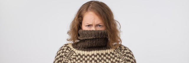 woman in brown and pale turtleneck with her face buried in the sweater looking at the camera