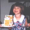 Photo of author as a child smiling in a dress holding a plate with a gingerbread house