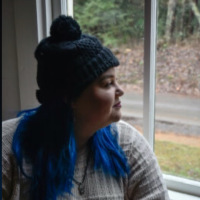 photo of author in a green hat with her blue hair out, looking outside the window. Wearing a pale sweater