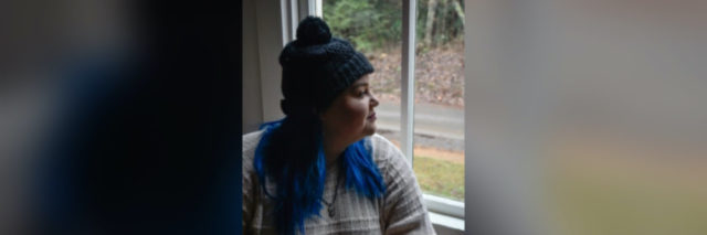 photo of author in a green hat with her blue hair out, looking outside the window. Wearing a pale sweater