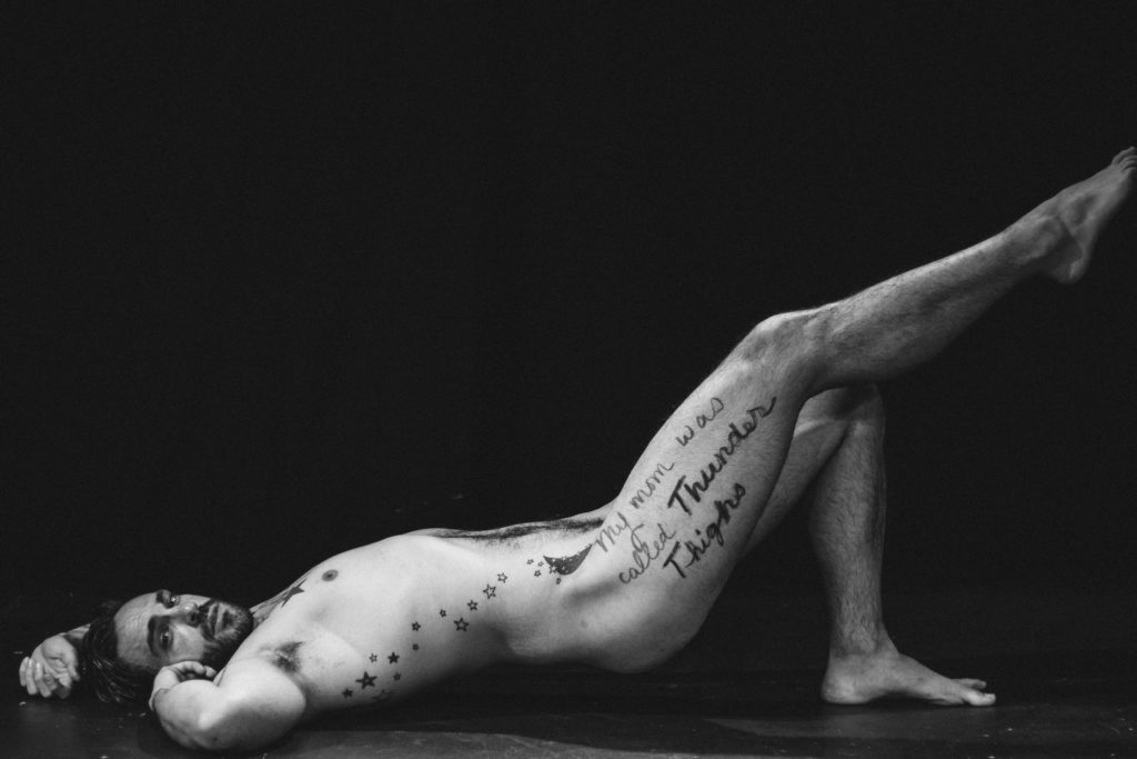 photo of man in black and white, lying nude with one leg raised. on his leg is written "my mom was called thunder thighs"