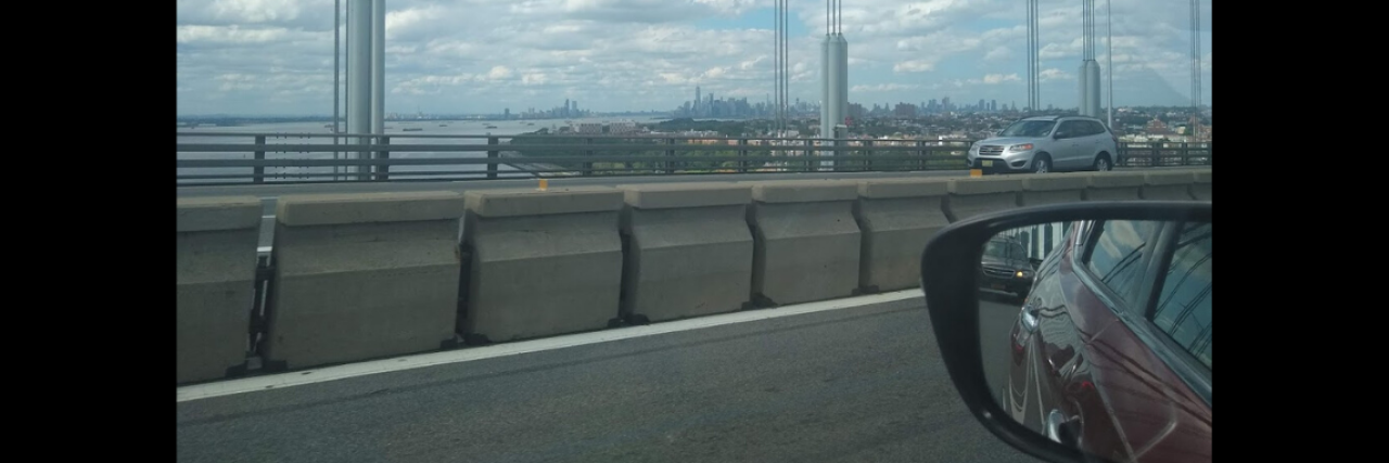 What Trauma Taught Me A scenic photo of a car driving over a bridge with clouds in the background.