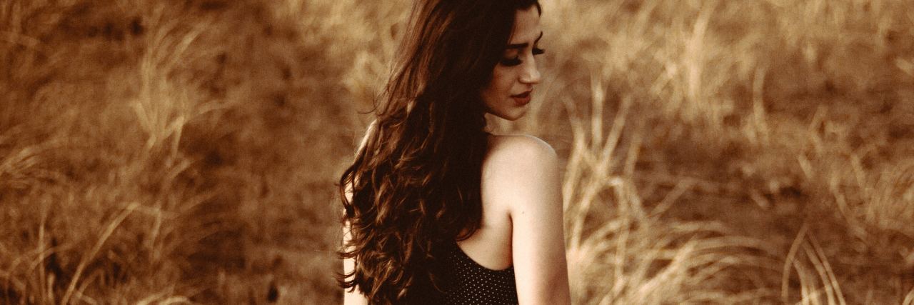 Bipolar Disorder Makes Me Feel Like a Burden on My Partner photo of woman standing in field with back to camera and looking over shoulder but down at the ground. in her hand is a hat.