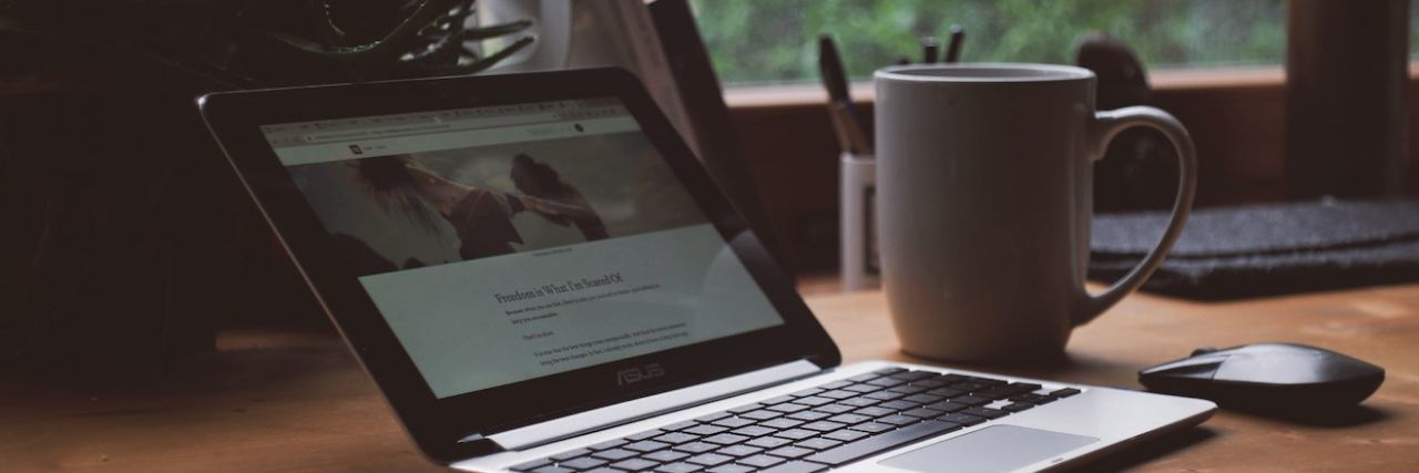 Work-From-Home Jobs for People With Disabilities and Chronic Illnesses Open laptop computer on a wooden desk by a window with a computer mouse and white coffee mug setting next to it