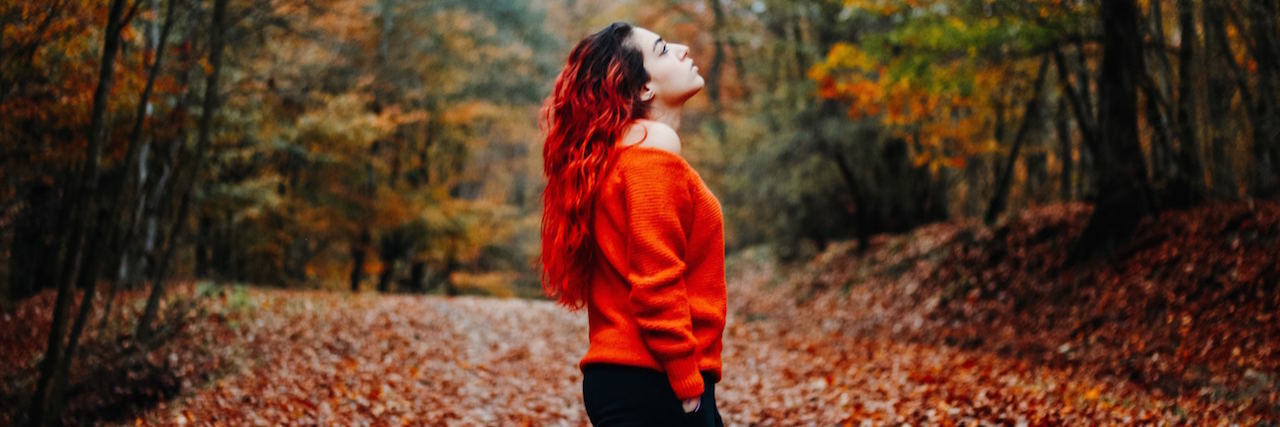 A woman with red hair in the woods looking up. The leaves are colorful - it's fall.