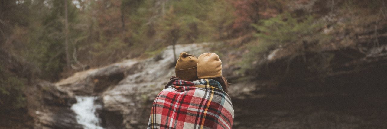2 girls wrapped in a blanket standing side by side with their backs to the camera in front of a lake