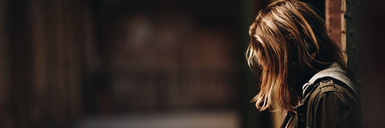 I'm Making Progress With My Depression, But I'm Still Not 'All Better' photo of a woman with her head down and hair covering her face standing in a dark hallway