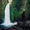 photo of young woman standing on rock by river and overlooking waterfall