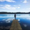 man in a flannel shirt and beanie standing on a long dock looking out into the blue water with clouds reflecting down