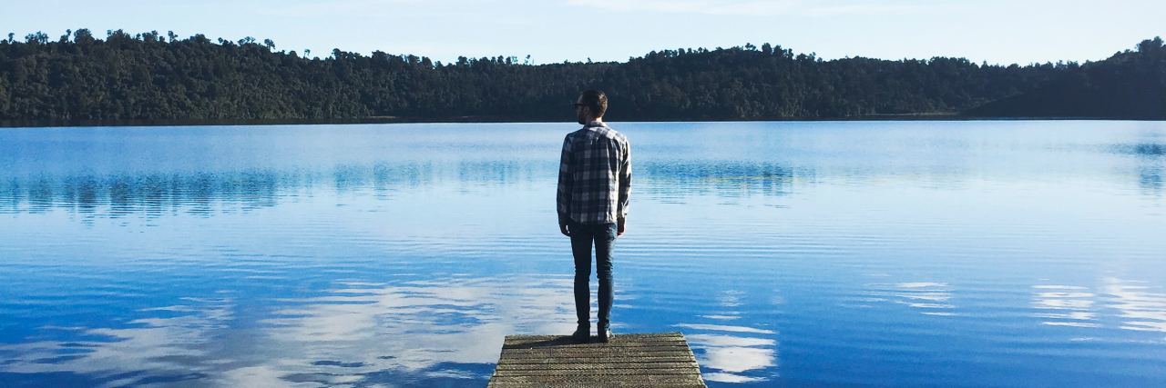man in a flannel shirt and beanie standing on a long dock looking out into the blue water with clouds reflecting down