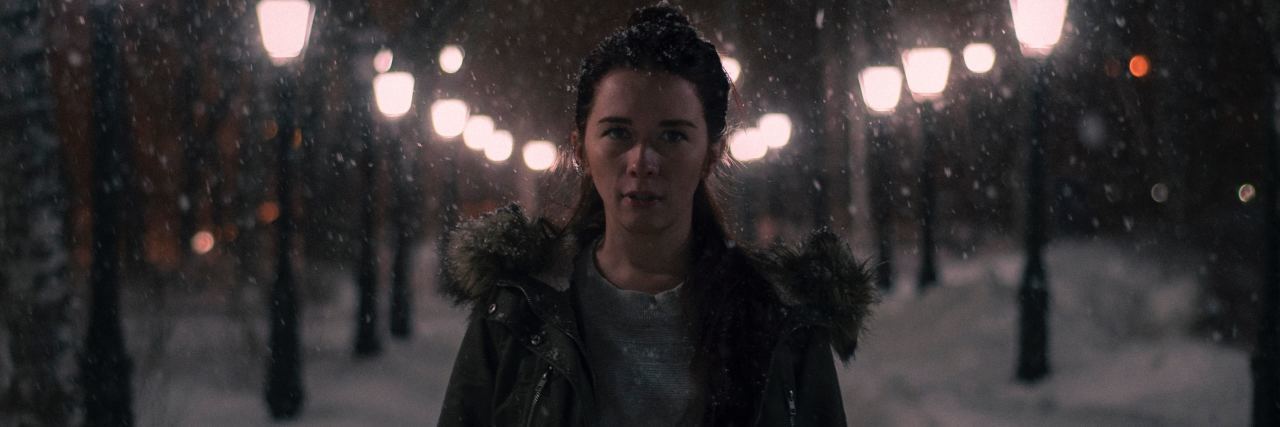 photo of woman at night standing on light-filled street in the snow, looking serious toward camera