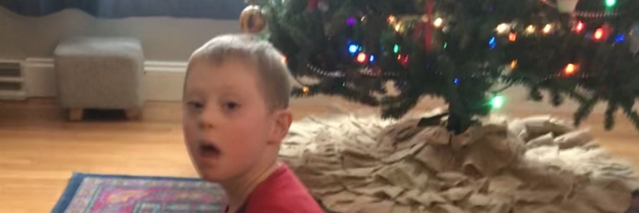 My Son With Down Syndrome Embodies All Things Christmas a boy in Christmas pajamas sitting near the Christmas tree