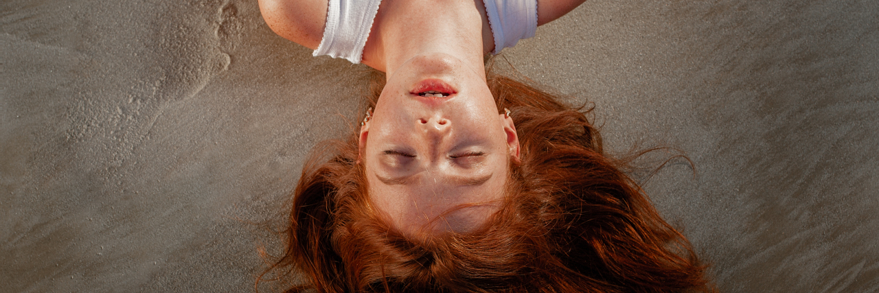 woman with red hair wearing a white shirt lying down on a beach with her eyes closed