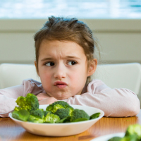 Unhappy girl eating spinach and broccoli at the table.