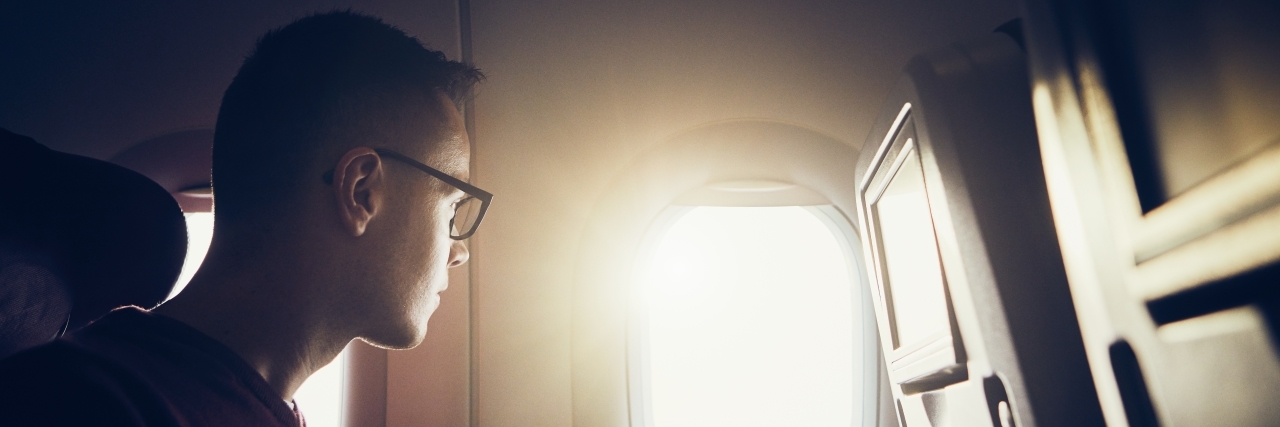 young man with glasses on an airplane in the window seat, looking outside the bright window