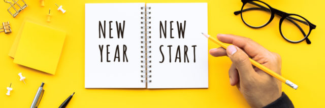 "New Year New Start" written on a notebook, person's hand with a pencil nearby
