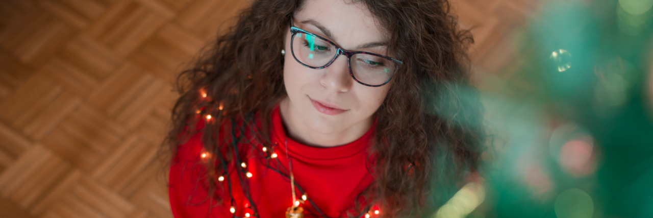 Why the Holidays Can Be Hard With Mental Illness photo of woman with dark hair and glasses sitting on floor beside Christmas tree looking thoughtful or pensive