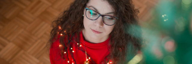photo of woman with dark hair and glasses sitting on floor beside Christmas tree looking thoughtful or pensive