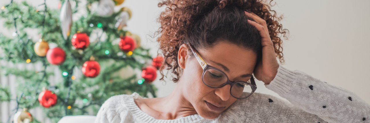 Missing Holiday Events Due to Chronic Illness Woman feeling sad during the holidays. She sits on a couch with a Christmas tree in the background.