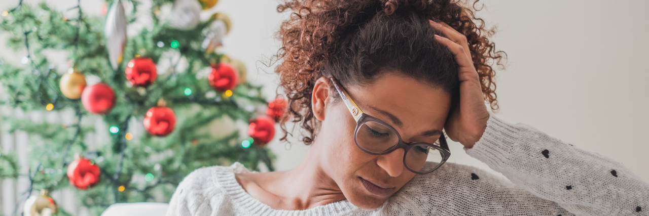 Woman feeling sad during the holidays. She sits on a couch with a Christmas tree in the background.