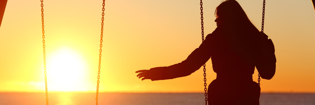 A woman sits alone in front of the sunset with an empty swing next to her.