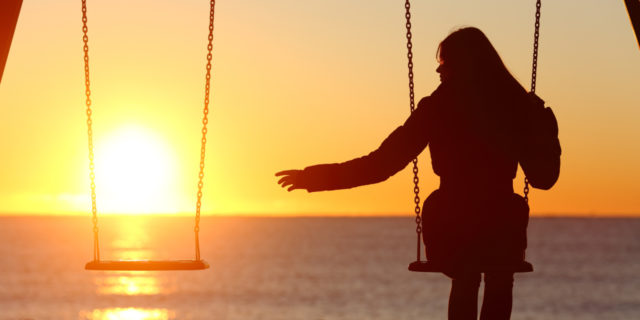 A woman sits alone in front of the sunset with an empty swing next to her.