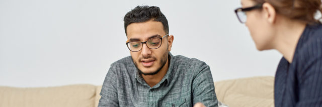 Young man wearing eyeglasses sitting on cozy sofa and discussing faced problem with highly professional psychologist during therapy session at cozy office