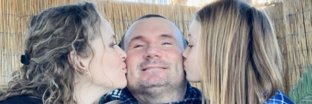 Jeff with his wife and daughter kissing him on each cheek.