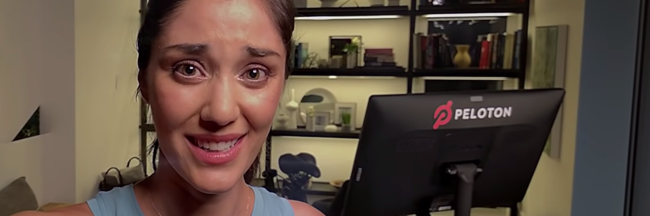 Take on Peloton Ad From a Health Coach With a Mental Illness screenshot from peloton exercise bike ad showing woman taking vlog with exercise bike behind her, looking in pain or tired