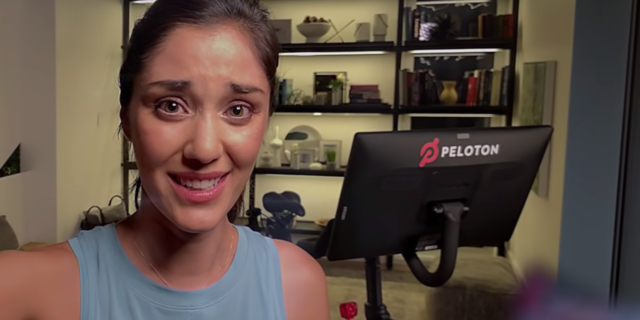 screenshot from peloton exercise bike ad showing woman taking vlog with exercise bike behind her, looking in pain or tired