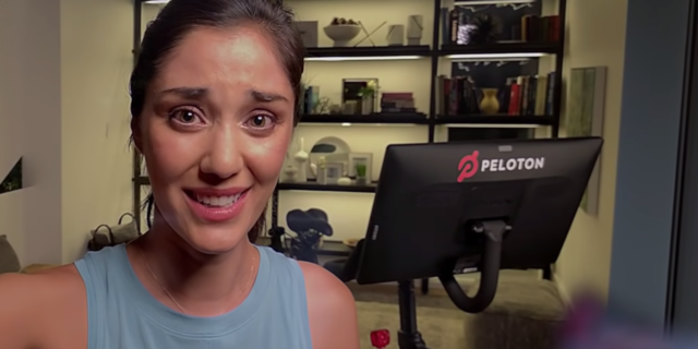 My Take on That Controversial Peloton Ad as a Health Coach With a Mental Illness screenshot from peloton exercise bike ad showing woman taking vlog with exercise bike behind her, looking in pain or tired