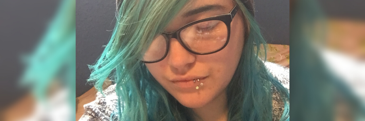 Taking a Selfie Every Day Changed My Relationship With My Body Image photo of contributor with blue hair, glasses and a hat which says "selfie", with eyes closed