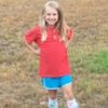a little girl smiling wearing a red shirt and blue shorts