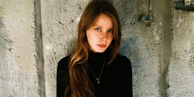photo of redheaded woman looking into camera with concerned expression, standing with her back against a concrete wall