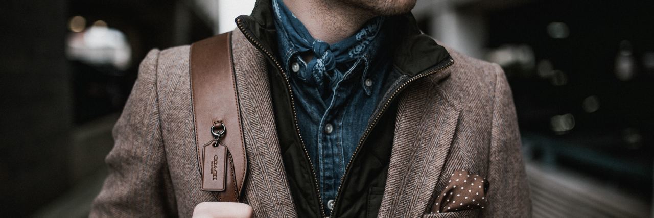 Leave Mental Illness 'Survival Mode' Behind When It Stops Serving You photo of bottom half of man's face down looking sideways in a brown suit jacket with his hand on a backpack strap