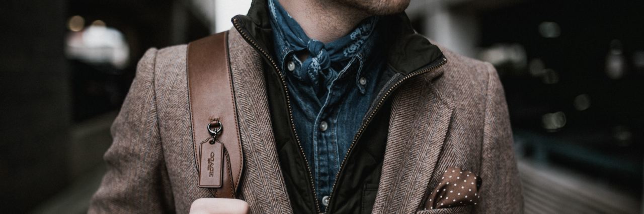 photo of bottom half of man's face down looking sideways in a brown suit jacket with his hand on a backpack strap