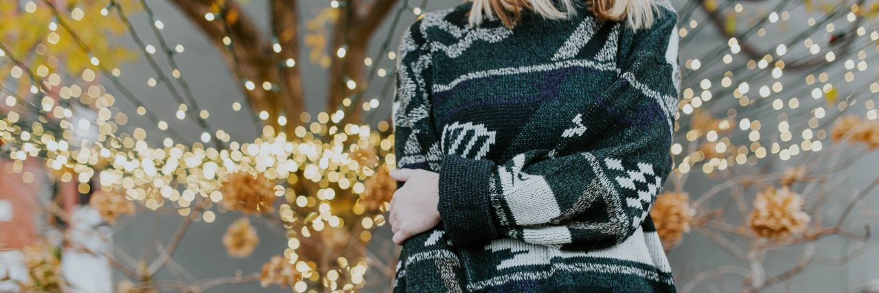 neck down picture of a woman in a green Christmas sweater standing in front of a lighted tree