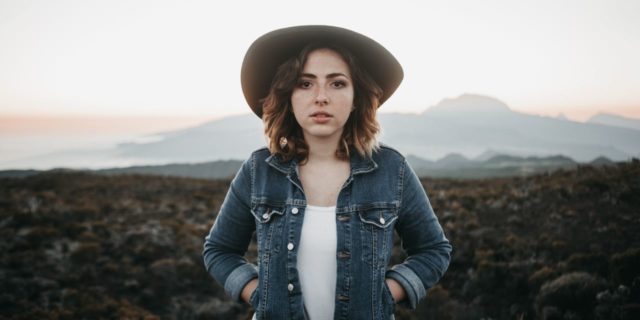 woman in a jean jacket and hat staring straight at camera with hands in her pockets