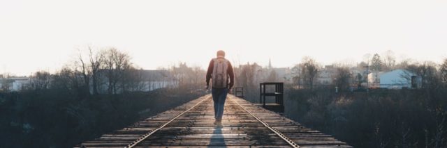 photo of a man on a railroad track walking with a backpack on toward the sunlight