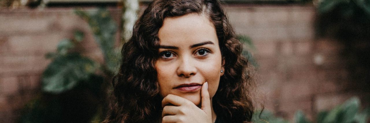 young woman with curly brown hair and a black shirt, staring at the camera with her hand on her chin