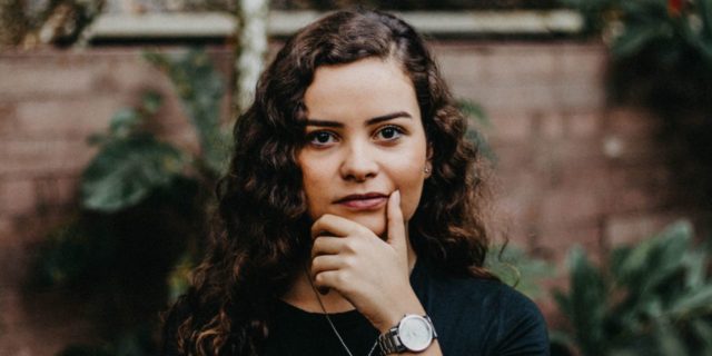 Dysphoric Mania Is Not 'Fun' young woman with curly brown hair and a black shirt, staring at the camera with her hand on her chin