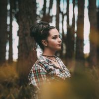 woman in a forest leaning against a tree with a straight face, hair in a bun and makeup on