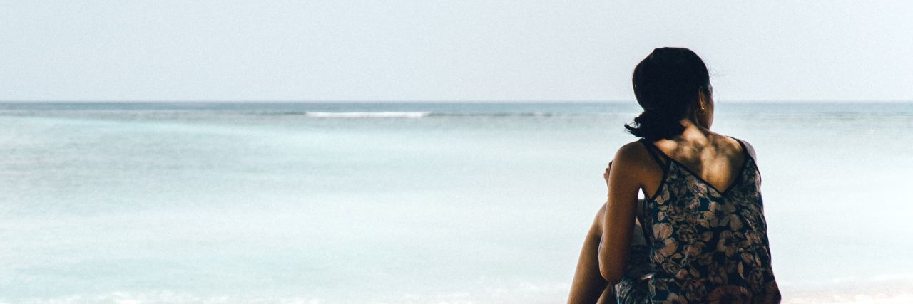 Vacations Can Be Stressful and Worrisome With Neurological Disease woman in a tank top sitting under a tree looking out into the ocean