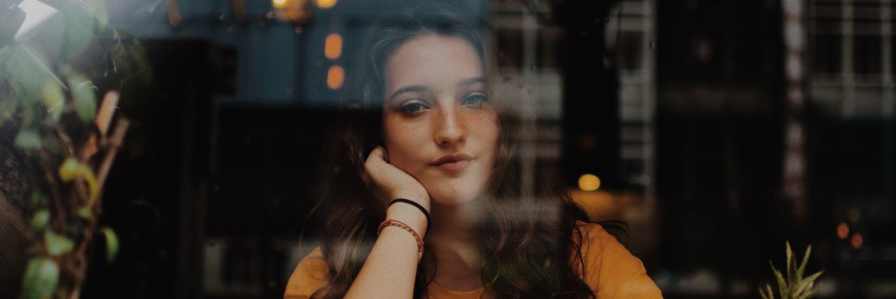 How I Manage 5 Emotional Burdens of Celiac Disease photo taken through a window of a young woman in a cafe looking outside