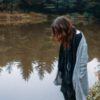 photo of woman standing in forest by lake or pond and looking into the water, her hair hiding her face