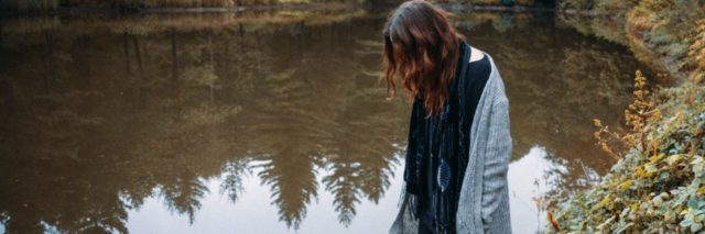 photo of woman standing in forest by lake or pond and looking into the water, her hair hiding her face