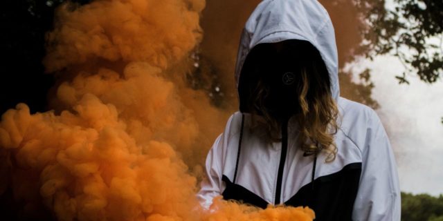 photo of woman with face covered by dark hair and hooded jacket, surrounded by orange smoke
