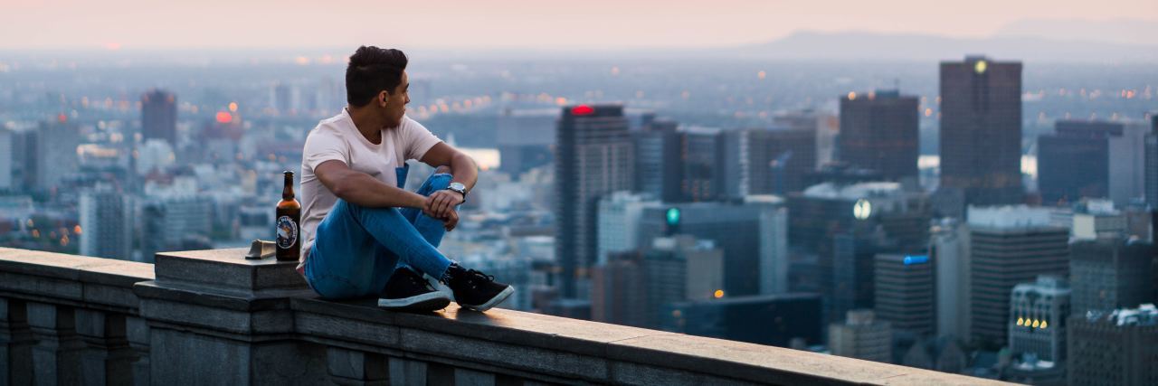young man sitting on the railing of a building with a beer next to him, overlooking a city