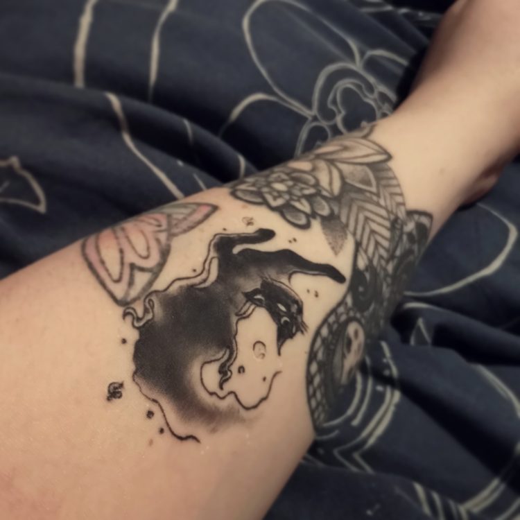 Foihnula shows the viewer a tattoo of a black cat with a cresent moon on their forehead.
